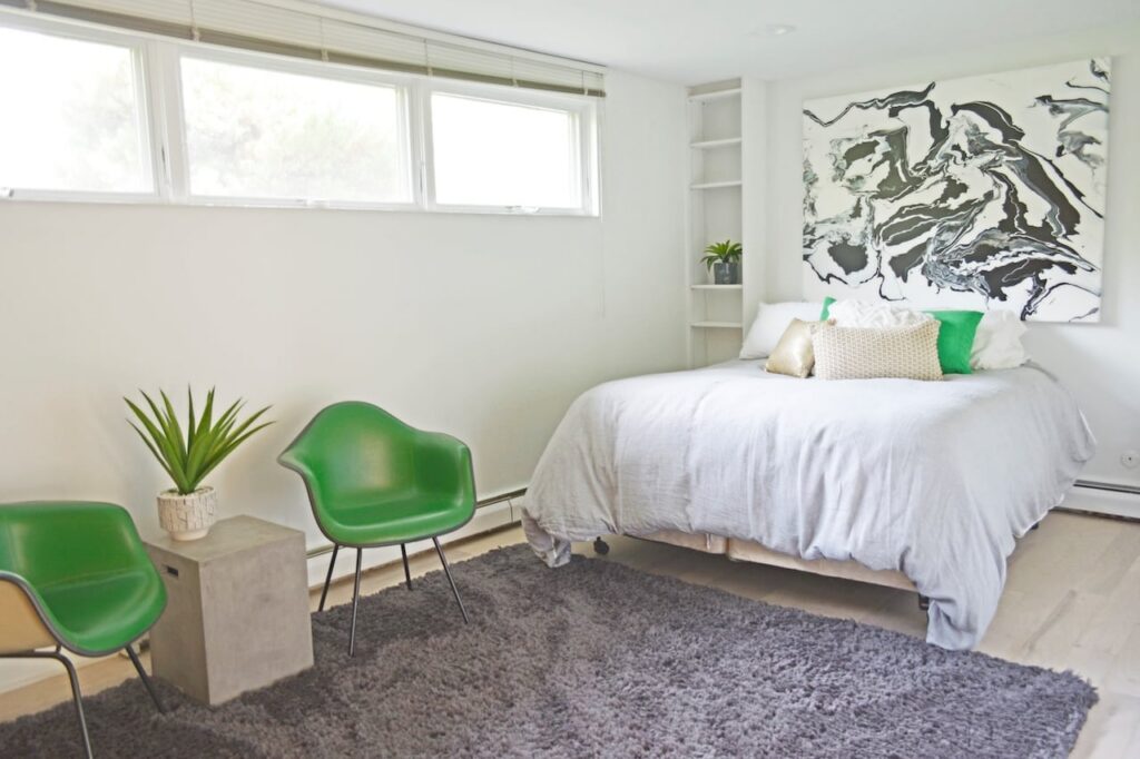 Minimalist bedroom with white bedding, green accent chairs, gray area rug, black and white abstract artwork, and horizontal window with natural light