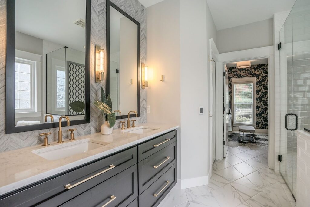 Modern luxury bathroom vanity with dual sinks, black frame mirrors, marble countertop, brass fixtures, and marble herringbone backsplash with glass shower enclosure