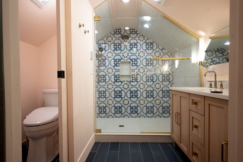 Master bathroom with blue and white geometric tile shower surround, gold-framed mirror, light wood vanity with brass fixtures, and dark tile floor