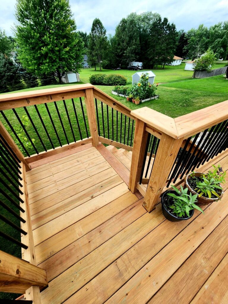 Wooden deck corner section with black vertical railings, wooden benches, and potted plants overlooking open green field