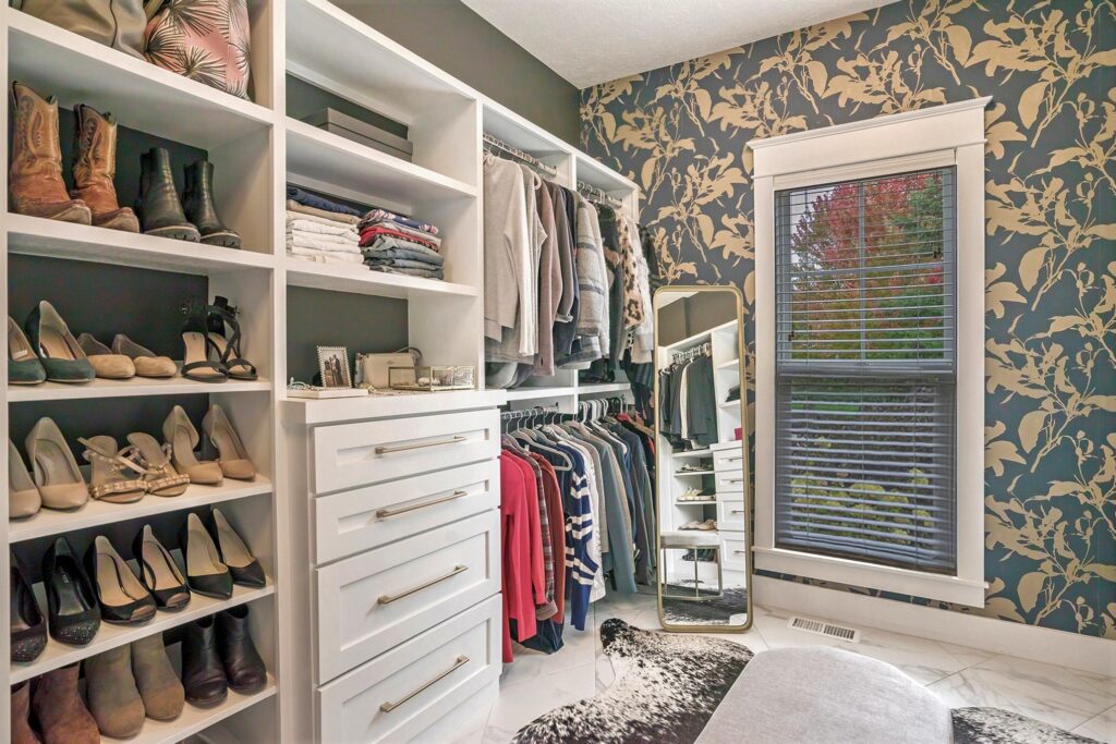 Luxury walk-in closet with white built-in shelving, shoe display, clothing rack, gold-framed mirror, and botanical patterned wallpaper